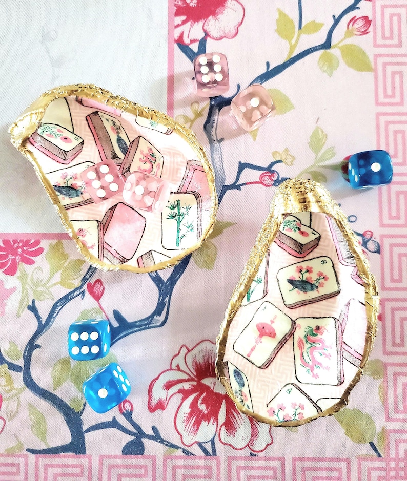 K&ouml;nnte beinhalten: Zwei dekorative Austernschalen mit goldenem Rand, gef&uuml;llt mit rosa und wei&szlig;en Mahjong-Steinen. Klare und blaue W&uuml;rfel sind verstreut, alles vor einem floral gemusterten Hintergrund.