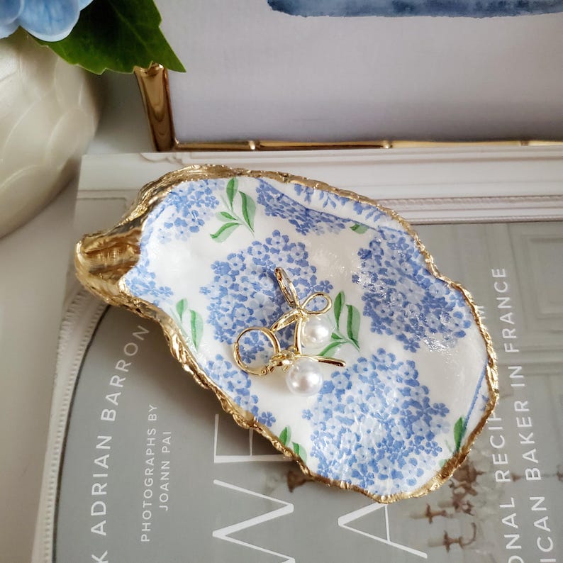 K&ouml;nnte beinhalten: Ein dekoratives Austernschalen-Schmucktablett mit goldenem Rand, verziert mit einem blau-wei&szlig;en Blumenmuster. Ein Paar goldene Ohrringe mit Perlenakzenten liegt darin. Das Tablett liegt auf einem Buch.