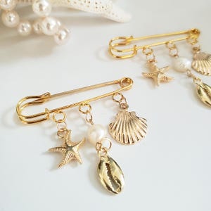 May include: Two gold-colored safety pins with dangling charms. Each pin features a pearl, starfish, seashell, and a shell charm. The pins are on a white surface, with a pearl necklace and a starfish in the background. The pins are likely for jewelry or fashion.