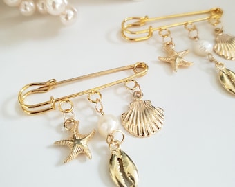 May include: Two gold-colored safety pins with dangling charms. Each pin features a pearl, starfish, seashell, and a shell charm. The pins are on a white surface, with a pearl necklace and a starfish in the background. The pins are likely for jewelry or fashion.