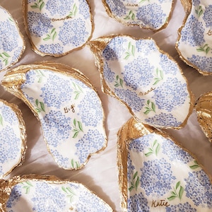 K&ouml;nnte beinhalten: Handbemalte Austernschalen mit blauen Hortensienbl&uuml;tenmustern und goldfarbenen R&auml;ndern. Jede Schale tr&auml;gt einen Namen in Schreibschrift. Die Schalen sind auf einer wei&szlig;en Oberfl&auml;che angeordnet.