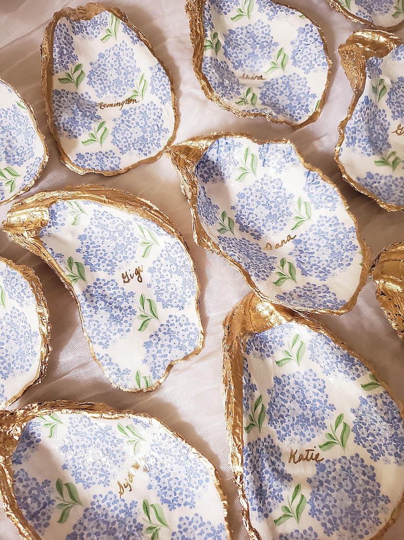 K&ouml;nnte beinhalten: Handbemalte Austernschalen mit blauen Hortensienbl&uuml;tenmustern und goldfarbenen R&auml;ndern. Jede Schale tr&auml;gt einen Namen in Schreibschrift. Die Schalen sind auf einer wei&szlig;en Oberfl&auml;che angeordnet.