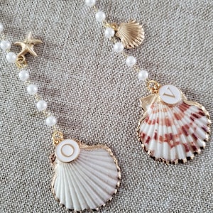 May include: A gold-toned necklace featuring seashell pendants, a starfish charm, and faux pearl beads. The seashell pendants have white and brown accents. The necklace is displayed on a textured, neutral-colored surface.