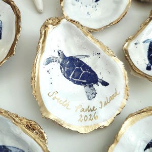 May include: White oyster shells with a gold rim, each featuring a blue sea turtle design. The words "South Padre Island 2026" are written in gold script on one shell. Decorative coastal theme.