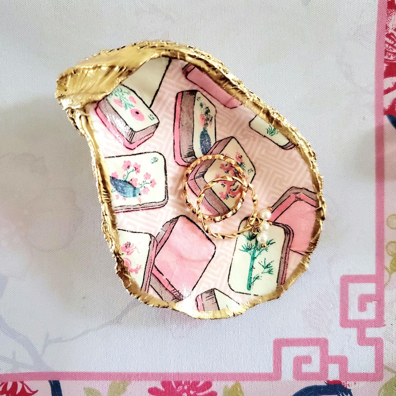 K&ouml;nnte beinhalten: Ein dekoratives Ringtablett aus Austernschale mit goldfarbenem Rand. Das Innere zeigt ein rosa-wei&szlig;es Muster mit floralen und geometrischen Motiven. Zwei goldene Ringe mit Perlenakzenten liegen darin.