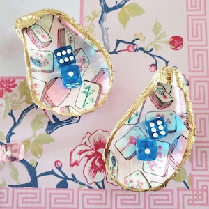 K&ouml;nnte beinhalten: Zwei dekorative Austernschalen mit goldfarbenen R&auml;ndern, die jeweils zwei blaue W&uuml;rfel enthalten. Die Schalen sind mit einem rosa-wei&szlig; gemusterten Stoff mit floralen und asiatisch inspirierten Motiven ausgekleidet. Ein einzelner rosa W&uuml;rfel ist sichtbar.