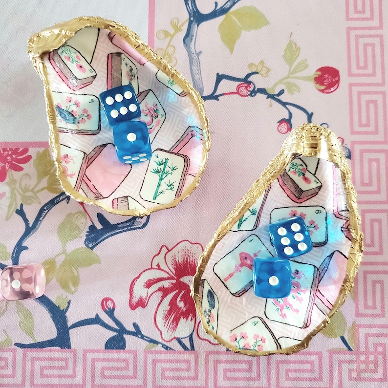K&ouml;nnte beinhalten: Zwei dekorative Austernschalen mit goldfarbenen R&auml;ndern, die jeweils zwei blaue W&uuml;rfel enthalten. Die Schalen sind mit einem rosa-wei&szlig; gemusterten Stoff mit floralen und asiatisch inspirierten Motiven ausgekleidet. Ein einzelner rosa W&uuml;rfel ist sichtbar.