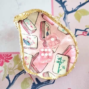 K&ouml;nnte beinhalten: Dekorative Austernschale, ausgekleidet mit rosa Stoff mit Mahjong-Fliesenmuster. Zwei rosa W&uuml;rfel mit wei&szlig;en Punkten liegen darin. Der Rand der Schale ist goldfarben. Dekoration.
