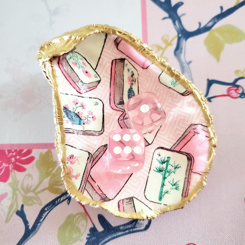 K&ouml;nnte beinhalten: Dekorative Austernschale, ausgekleidet mit rosa Stoff mit Mahjong-Fliesenmuster. Zwei rosa W&uuml;rfel mit wei&szlig;en Punkten liegen darin. Der Rand der Schale ist goldfarben. Dekoration.
