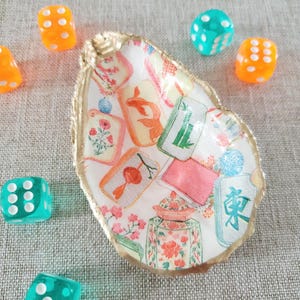 May include: A decorative oyster shell dish with a gold-painted rim, featuring a decoupage pattern of colorful mahjong tiles, floral motifs, and a ginger jar. The dish is surrounded by several translucent orange and teal six-sided dice.