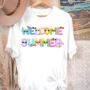 May include: White t-shirt with the words "WELCOME SUMMER" in colorful bubble letters. The letters are decorated with sunglasses, watermelon slices, lemons, and stars. A tan hat hangs on the hanger above the shirt.