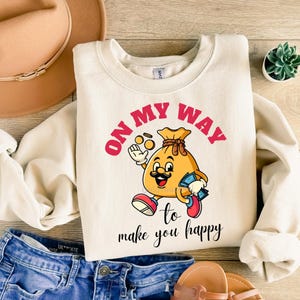 Peut inclure: Sweat-shirt crème avec l'inscription "ON MY WAY" en rose et un personnage de sac d'argent de dessin animé. Le personnage a une moustache, des chaussures rouges et tient des pièces et de l'argent. Le texte "to make you happy" est en dessous.