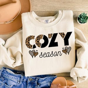 以下が含まれることがあります： 黒とヒョウ柄の「COZY season」という文字が入ったクリーム色のスウェットシャツ。ヒョウ柄と黒いハートの2つのハートの形が「season」という言葉の下にあります。