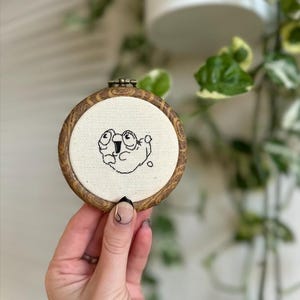 Puede incluir: Un pequeño bastidor de bordado redondo con un marco de madera. La tela blanquecina del interior presenta un dibujo lineal negro caprichoso de un personaje de dibujos animados con ojos grandes. El bastidor se sostiene en una mano, con un fondo borroso de vegetación.