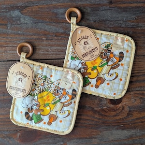 May include: Two vintage kitchen pot holders with a raccoon chef design. The pot holders have a yellow and white plaid pattern with a wooden ring for hanging. Each pot holder has a tag that says "Kitchen Compliments".