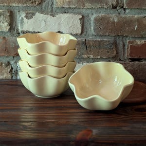 May include: A set of five pale yellow ceramic bowls with a distinct wavy, scalloped rim design. Four of the bowls are stacked vertically in a column, while one individual bowl is positioned to the right of the stack on a dark wooden surface.
