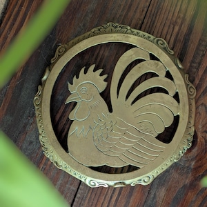 May include: A brass-colored, circular trivet featuring a detailed rooster design within a decorative frame. The rooster is depicted in profile, with intricate feather and comb details. The trivet is set against a dark wood surface.