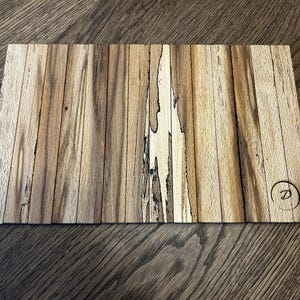 Puede incluir: Tabla de cortar rectangular de madera hecha de tiras verticales de madera clara y oscura. La veta de la madera es visible, con un logotipo circular con la letra "D" grabada en la esquina inferior derecha. Sobre una superficie de madera.