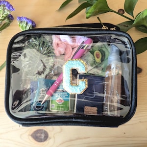 May include: A clear, black-trimmed makeup bag with a zipper closure. The bag contains various items, including a pink scrunchie, a pink pen, a clear bottle with a gold lid, and a blue letter "C" patch.