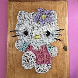 May include: String art of a cartoon cat character on a wooden board. The cat is white with black eyes and whiskers, wearing a pink top and a purple flower. The board is a light brown color.