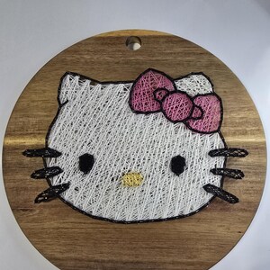 May include: A round wooden plaque featuring a string art design of Hello Kitty. The design includes a pink bow, black whiskers, and black eyes. The background is a light brown wood with a hole at the top for hanging.