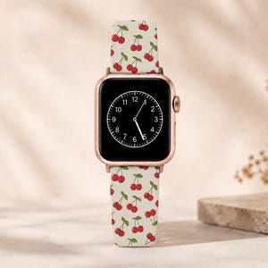 May include: A rose gold smartwatch with a black screen displaying the time. The watch band is light beige with a pattern of red cherries and green leaves. The watch is displayed on a neutral background.