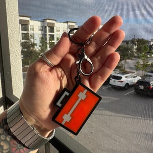May include: A rectangular keychain with a black border and a bright orange center. A white vertical stripe and two horizontal white stripes are in the center. The keychain has a black keyring and clasp.