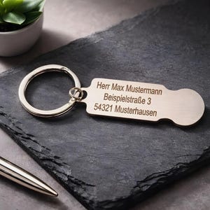 Könnte beinhalten: Ein silberfarbener Metall-Schlüsselanhänger mit einem rechteckigen Anhänger, auf dem der Text "Herr Max Mustermann, Beispielstraße 3, 54321 Musterhausen" eingraviert ist. Der Schlüsselanhänger liegt auf einer dunkelgrauen Oberfläche.