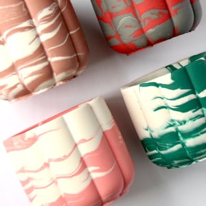 May include: Four small, square planters with a ribbed design and marbled patterns. Colors include shades of pink, red, brown, green, and white. The planters are arranged on a white background.