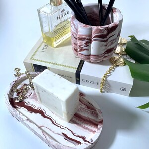 May include: A decorative vanity set featuring a white and burgundy marbled tray with a bar of soap, a matching pen holder with makeup brushes, and a bottle of perfume. A book and gold chain are also present.
