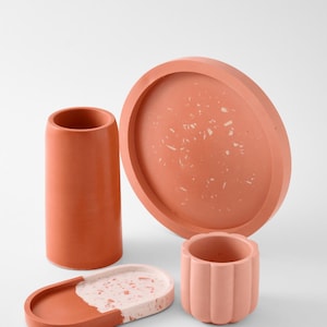 Può includere: Una collezione di articoli per l'arredamento della casa in cemento color terracotta. Il set comprende un vaso cilindrico alto, un vassoio rotondo con un design maculato, un piccolo vaso smerlato e un vassoio ovale con un design maculato.