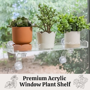 May include: A clear acrylic window plant shelf with three potted plants. The shelf is attached to the window with suction cups. The plants are in terracotta, white, and beige pots. The text "Premium Acrylic Window Plant Shelf" is at the bottom.