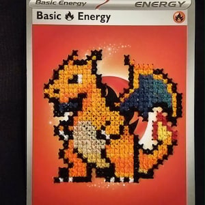 May include: A rectangular trading card featuring a red background with the text Basic Energy at the top. In the center, a pixelated, cross-stitched orange dragon character with blue wings is displayed. The card includes a small fire symbol in the corner.