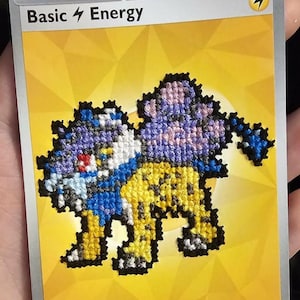 Op de afbeelding: Een Basic Energy Pokémon-ruilkaart met een gele achtergrond en een bliksemsymbool. In het midden staat een kruissteekontwerp van een viervoetig wezen met een geel lichaam, blauwe accenten en een paarse, wolkachtige vorm op de rug.