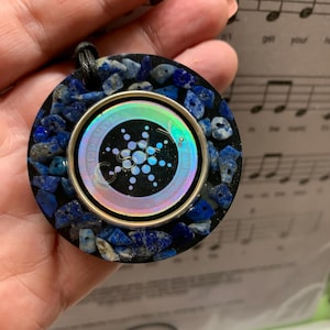 May include: A round pendant with a black base, encircled by blue stones. The center features a silver-rimmed iridescent design with the text "QUANTUM-SCIENTIFIC". A black cord is attached for wearing.