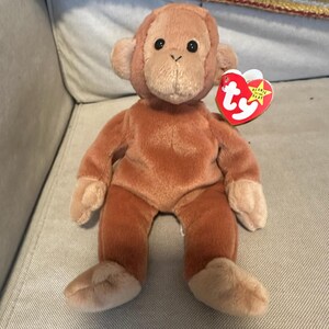 Puede incluir: Un mono de peluche marrón con una etiqueta Ty roja que dice "Beanie Babies" y "Ty".
