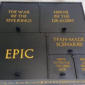 May include: A black wooden box with several compartments, each labelled with gold text. The box is divided into sections with phrases such as "The War of the Five Kings", "House of the Dragon", and "Epic". It appears to be a storage solution for a board game.