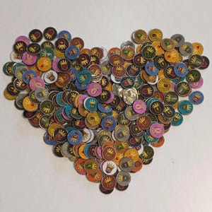 Könnte beinhalten: Eine herzförmige Anordnung von bunten runden Spielsteinen mit verschiedenen Designs, darunter Kronen, Schilde und andere Symbole. Die Spielsteine sind aus Metall und haben eine glänzende Oberfläche.