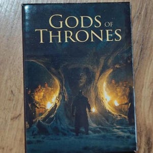 以下が含まれることがあります： 暗い青色の背景に金色の文字で「Gods of Thrones」と書かれた本の表紙。表紙には、後ろからオレンジ色の光が放たれている大きな木のような構造物の前に立っている、暗くて影のある人物が描かれている。