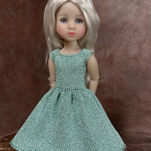 Op de afbeelding: Een pop met lang, steil, lichtblond haar en blauwe ogen, gekleed in een kortmouwige, getextureerde, saliegroene gebreide jurk met een aansluitend lijfje en een uitlopende rok, gecombineerd met licht mintgroene sandalen met kleine bloemversieringen.