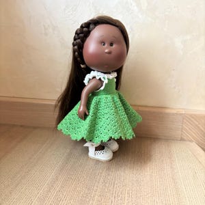 May include: A handmade doll with dark skin, brown hair in a braid, and a green crocheted dress. The doll wears white shoes with small cut-out details. The dress has a white lace collar. The doll is standing on a wooden surface.