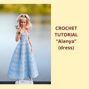 Op de afbeelding: Een pop draagt een lichtblauwe gehaakte jurk met een halternek en een wijde rok. De jurk maakt deel uit van een haak tutorial genaamd "Alanya". De pop heeft blond haar en staat op een witte basis. De achtergrond is wazig.