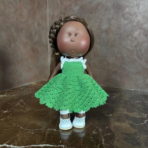 May include: A handmade doll with brown skin, brown hair in a braid, and a round face. The doll wears a green crocheted dress with white trim and white shoes. The doll is standing on a brown surface.