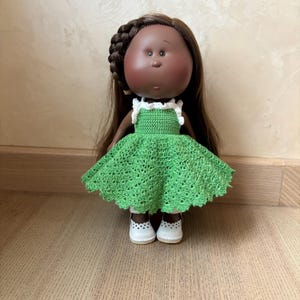 May include: A handmade doll with dark skin, brown hair, and a green crocheted dress with white trim. The doll is wearing white shoes with small cut-out details. The doll's hair is styled with a braid.