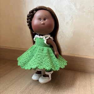 May include: A handcrafted doll with brown hair, a braided side, and a green crocheted dress with white trim. The doll wears white shoes with cut-out details. The doll has a dark complexion and is standing on a wooden surface.
