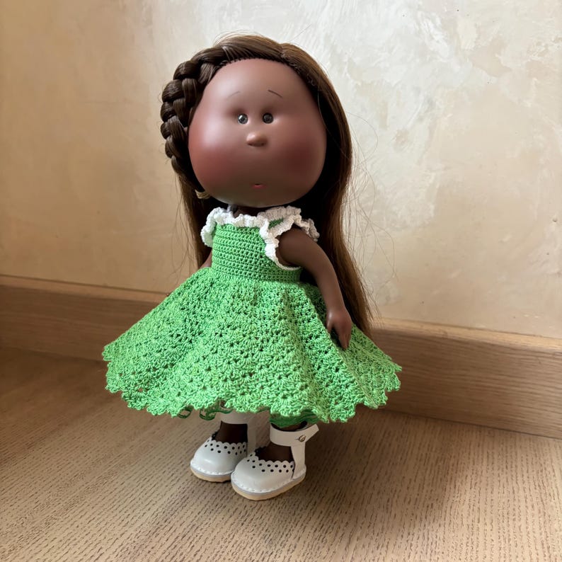 May include: A handcrafted doll with brown hair, a braided side, and a green crocheted dress with white trim. The doll wears white shoes with cut-out details. The doll has a dark complexion and is standing on a wooden surface.