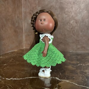 May include: A handmade doll with brown skin, brown hair in a braid, and a green crocheted dress with white trim. The doll wears white shoes and has a sweet expression. The doll is a charming handmade item.
