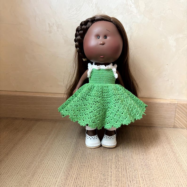 May include: A handmade doll with brown skin, brown hair, and a green crocheted dress. The doll is wearing white shoes with black polka dots. The doll's hair is styled with a braid on top of the head.