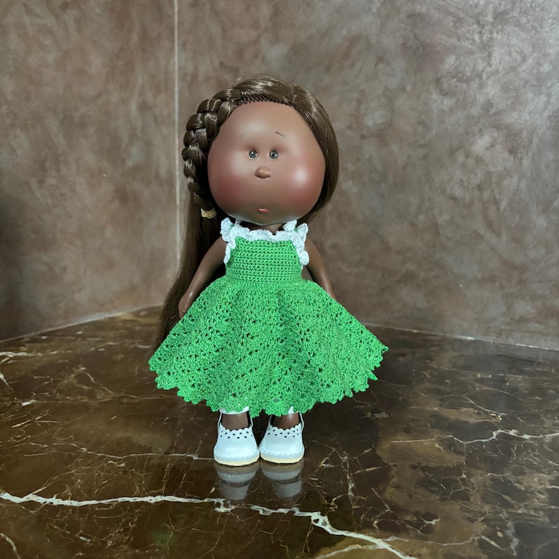 May include: A handmade doll with brown hair styled in a braid and long strands. The doll has a dark complexion and is wearing a green crocheted dress with white trim and white shoes. The doll is standing on a reflective surface.