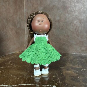 May include: A handcrafted doll with brown hair styled in a braid. The doll wears a bright green crocheted dress with white trim and white shoes. The doll has a round face with rosy cheeks and a small nose.